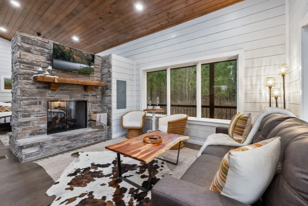 Settle into this sunroom where the stone fireplace glows, the forest wraps around you through those massive windows, and you realize you've found the perfect spot to do absolutely nothing but exist.