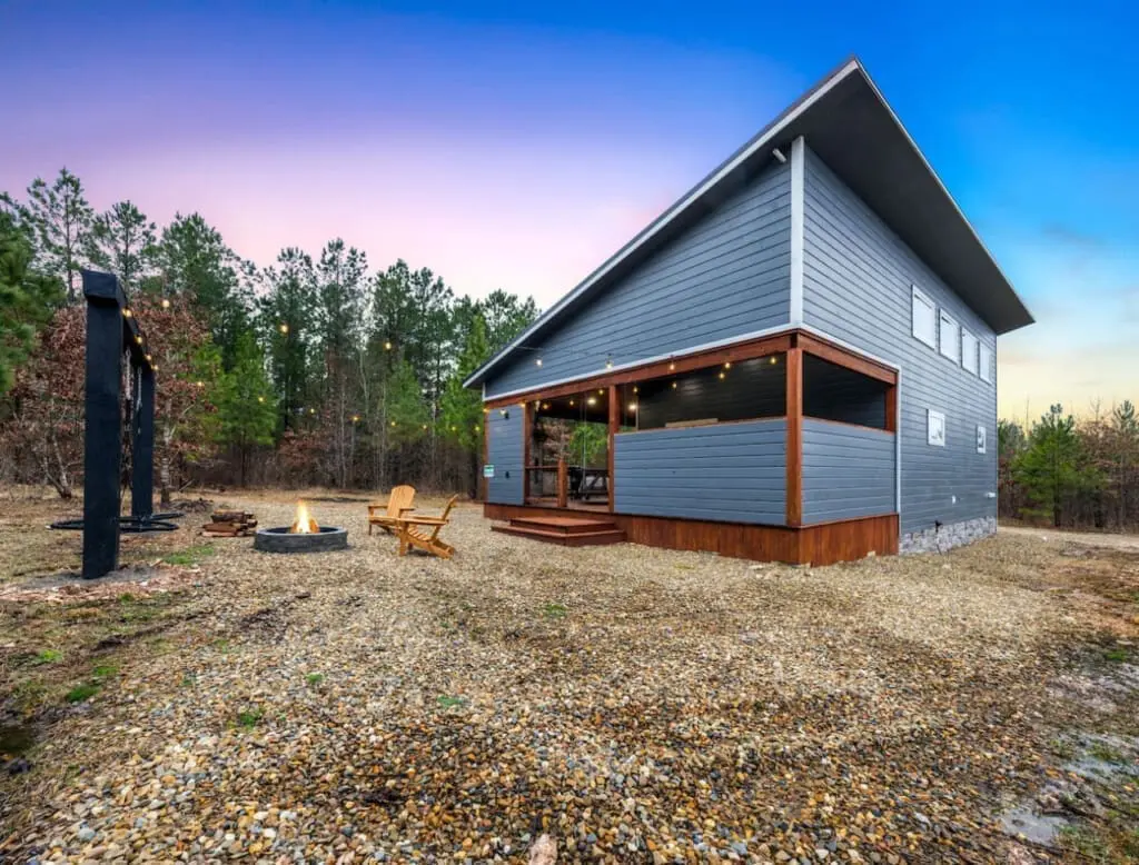 Watch the sky shift through every shade of blue and purple as the sun sets over your private forest escape, fire pit glowing warm below and that modern cabin silhouette looking like it was plucked straight from your Pinterest board.