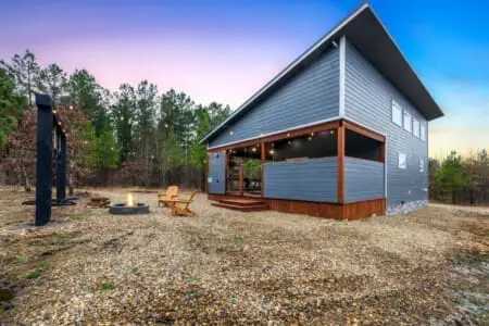 Watch the sky shift through every shade of blue and purple as the sun sets over your private forest escape, fire pit glowing warm below and that modern cabin silhouette looking like it was plucked straight from your Pinterest board.