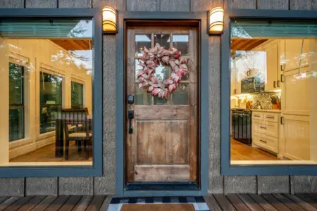 This entryway basically sets the tone the second you arrive—rustic wood door, those welcoming sconces glowing on either side, and those huge windows framing the warm interiors that make you feel like you're stepping into somewhere special.