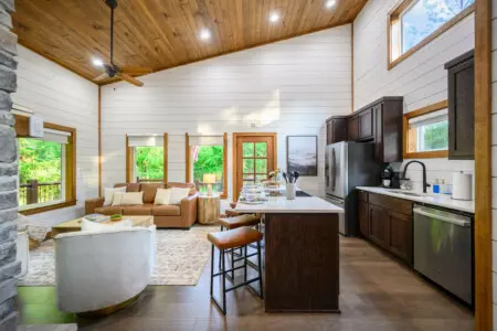 The whole vibe of this place just *works*—white shiplap walls, warm wood ceilings, a kitchen island that begs for conversation, and those floor-to-ceiling windows bringing the forest right into your gathering space.