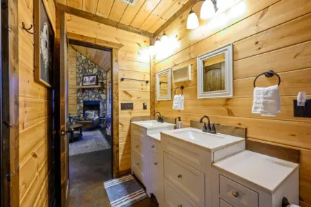 This gorgeous log cabin bathroom features dual vanities, stone fireplace views, and rustic wood finishes throughout.