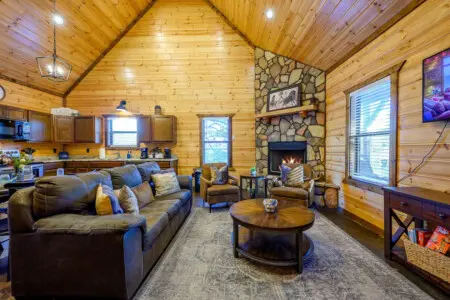 This spacious log cabin living room features a stone fireplace, cozy leather seating, and plenty of natural light from every angle.