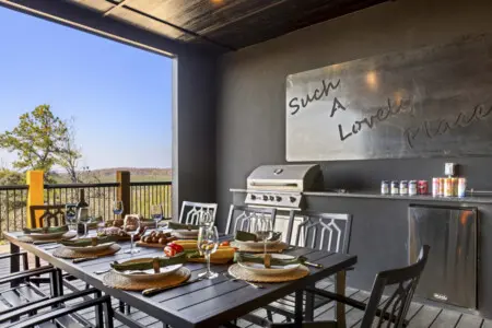 Outdoor kitchen with a grill, built-in fridge, and a table for eight overlooking the trees.