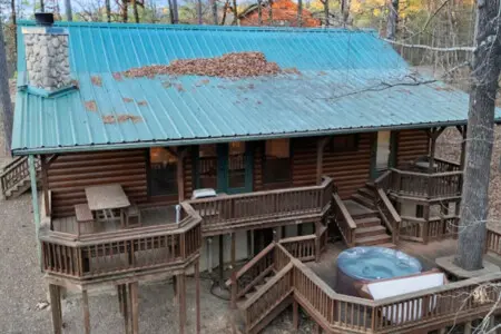 This gorgeous log cabin features wraparound decks, a hot tub, and a turquoise metal roof nestled in the woods.