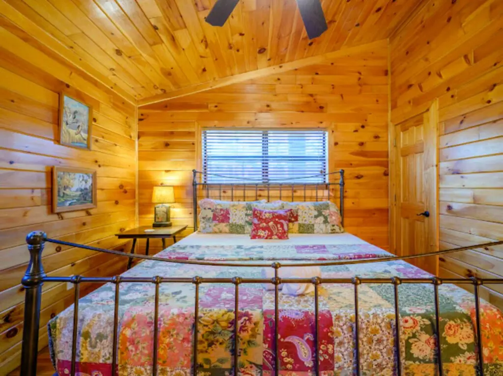 Cozy log cabin bedroom with warm wood throughout, a colorful quilt-covered bed, and plenty of natural light from the big window.