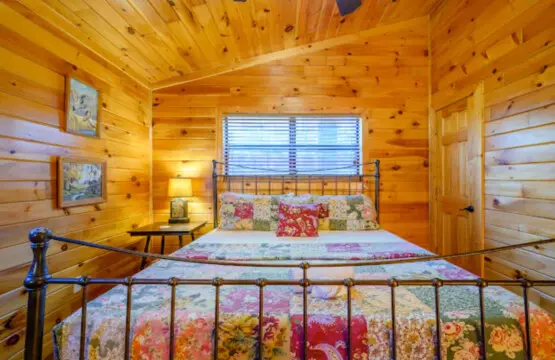 Cozy log cabin bedroom with warm wood throughout, a colorful quilt-covered bed, and plenty of natural light from the big window.