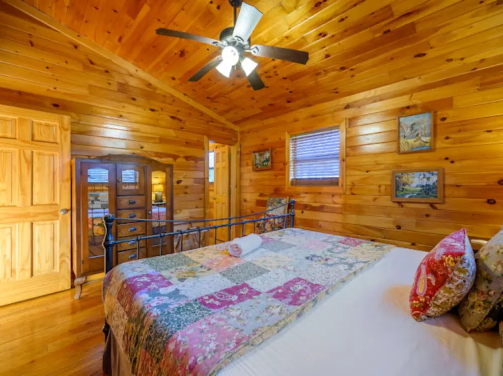 This cozy log cabin bedroom features warm wood throughout, a comfortable bed, and a ceiling fan to keep things cool.