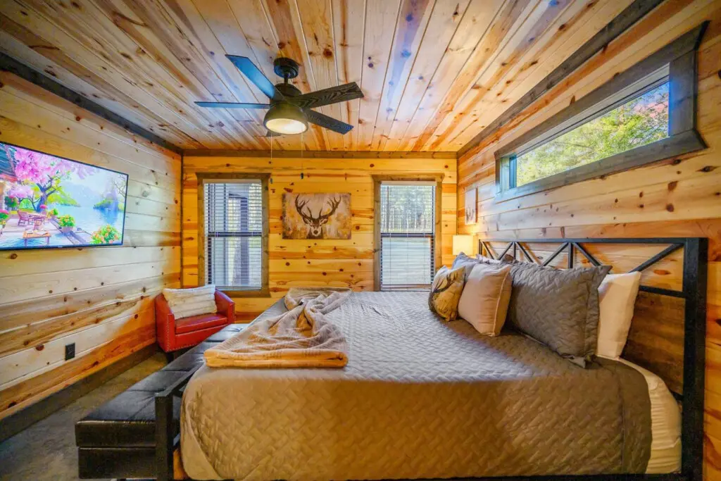 This log cabin bedroom is basically the ultimate cozy retreat with those warm wood walls surrounding you, a spacious bed loaded with pillows, a ceiling fan overhead, and those big windows framing the forest while a TV and that red leather chair give you p