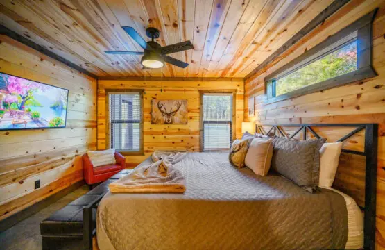 This log cabin bedroom is basically the ultimate cozy retreat with those warm wood walls surrounding you, a spacious bed loaded with pillows, a ceiling fan overhead, and those big windows framing the forest while a TV and that red leather chair give you p