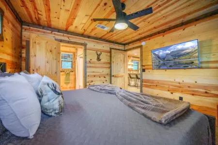 This master bedroom totally nails the cabin aesthetic with those warm knotty pine walls, a spacious bed setup, an en-suite bathroom, and even a TV playing mountain scenery in case you need a backup view.