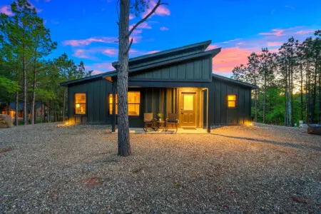 This sleek modern cabin feels like your own private retreat tucked into the pines, with that warm glow spilling out onto the gravel courtyard just as the sky turns that perfect shade of blue and pink.