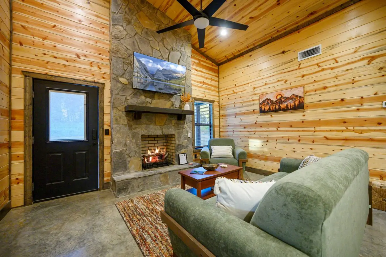 This cozy log cabin living room has everything you need to settle in—a stone fireplace with a real fire going, comfy green chairs, and even a mountain view on the TV if you need a backup when you're not gazing out the actual windows.