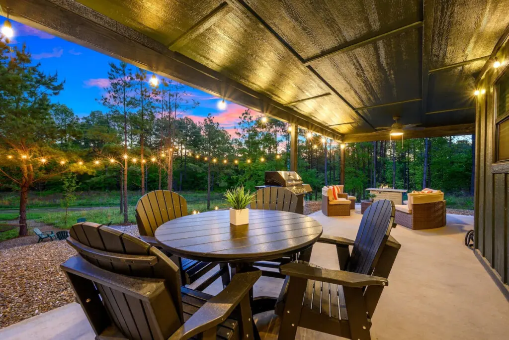 This covered patio setup is giving major entertaining energy with that spacious dining table, lounge seating, and a built-in grill all wrapped up under warm lighting while the forest melts into that gorgeous twilight sky.