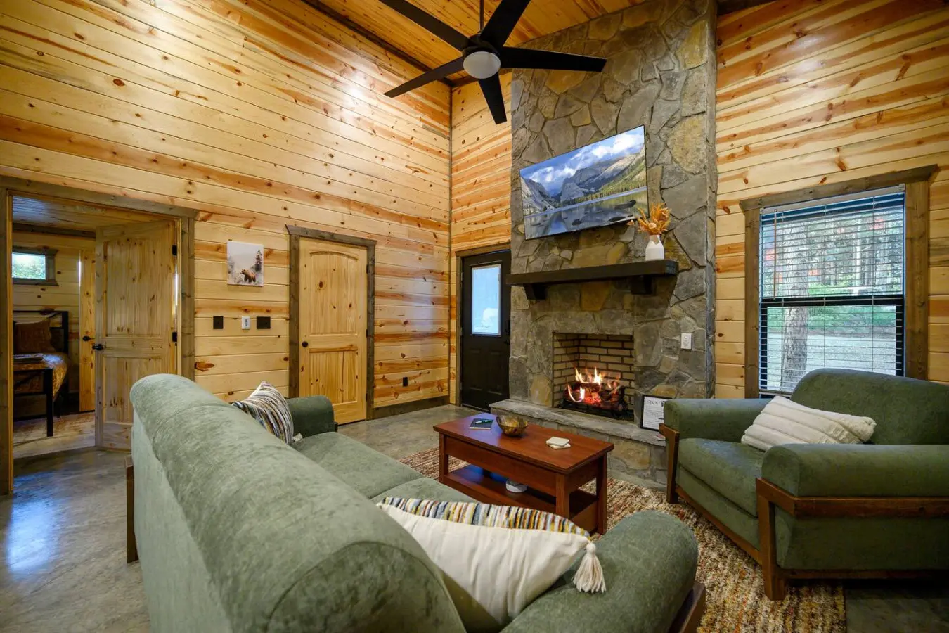This log cabin living room is basically the perfect setup for hanging out—you've got the stone fireplace doing its thing, those comfy green chairs, a TV to keep you entertained, and all that natural wood making it feel like you're genuinely tucked away in