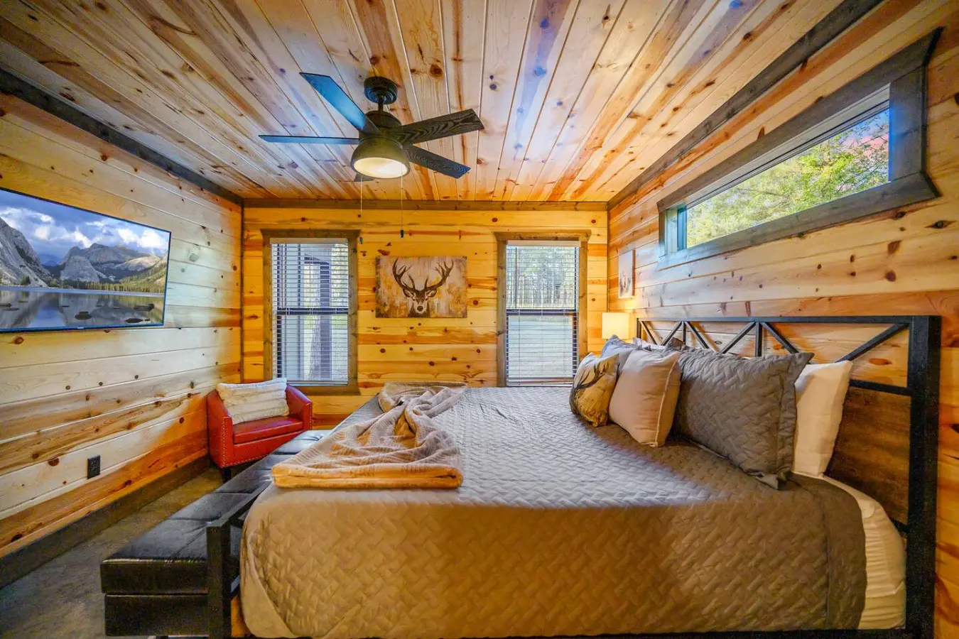 This master bedroom is basically the ultimate cabin escape with those knotty pine walls, a killer bed setup, a ceiling fan to keep things comfortable, and windows framing the trees while a TV and cozy seating keep you entertained on lazy mornings.