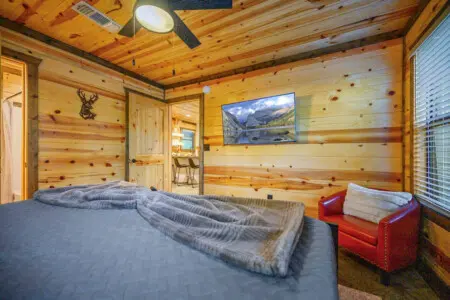 This log cabin master bedroom has all the cozy-retreat energy—knotty pine walls, a plush bed piled with blankets, a flat-screen showing mountain views, and that red leather chair in the corner for when you just want to settle in and watch the world go by.