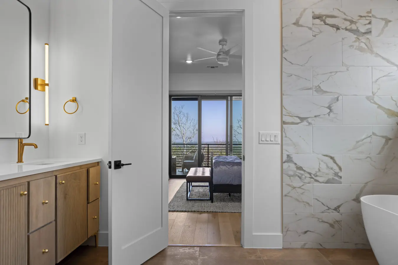 The master bath has marble tile, gold fixtures, and a soaking tub with a view straight through to the bedroom balcony.