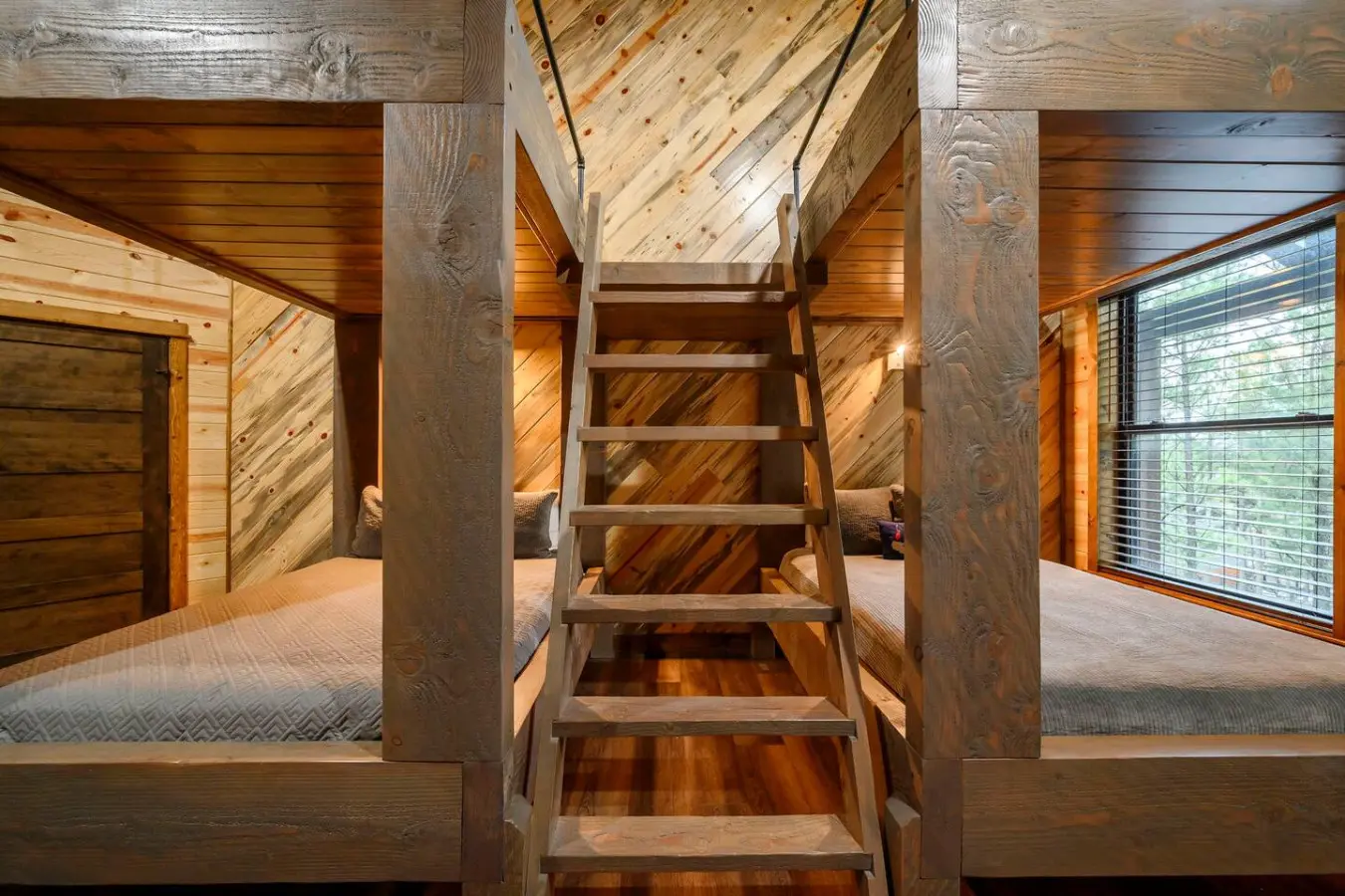 This cozy loft bedroom features twin beds tucked under soaring timber ceilings with a rustic wooden ladder connecting the two sleeping spaces.