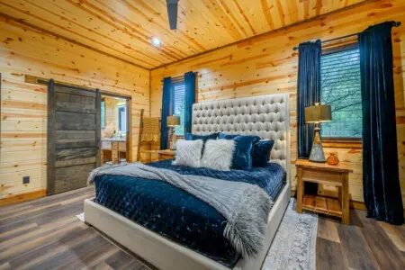 This stunning log cabin bedroom features a plush tufted headboard, cozy wood walls, and elegant teal curtains framing the forest views.