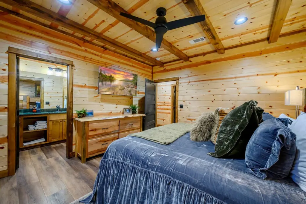 This cozy log cabin bedroom features exposed wooden beams, a ceiling fan, and an ensuite bathroom with plenty of natural light.