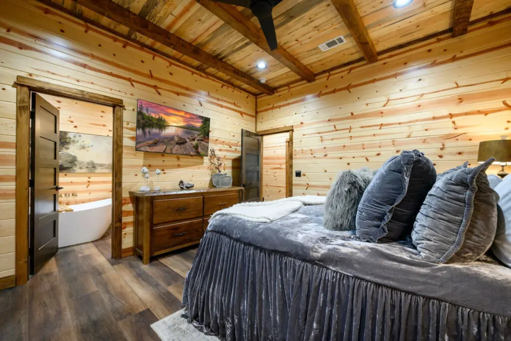 This cozy log cabin bedroom features soaring wood beams, an ensuite bathroom, and stunning landscape artwork above the dresser.