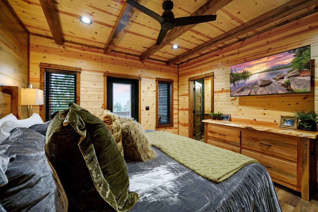 This spacious log cabin bedroom features exposed beams, multiple doors to nature views, and plenty of natural light.