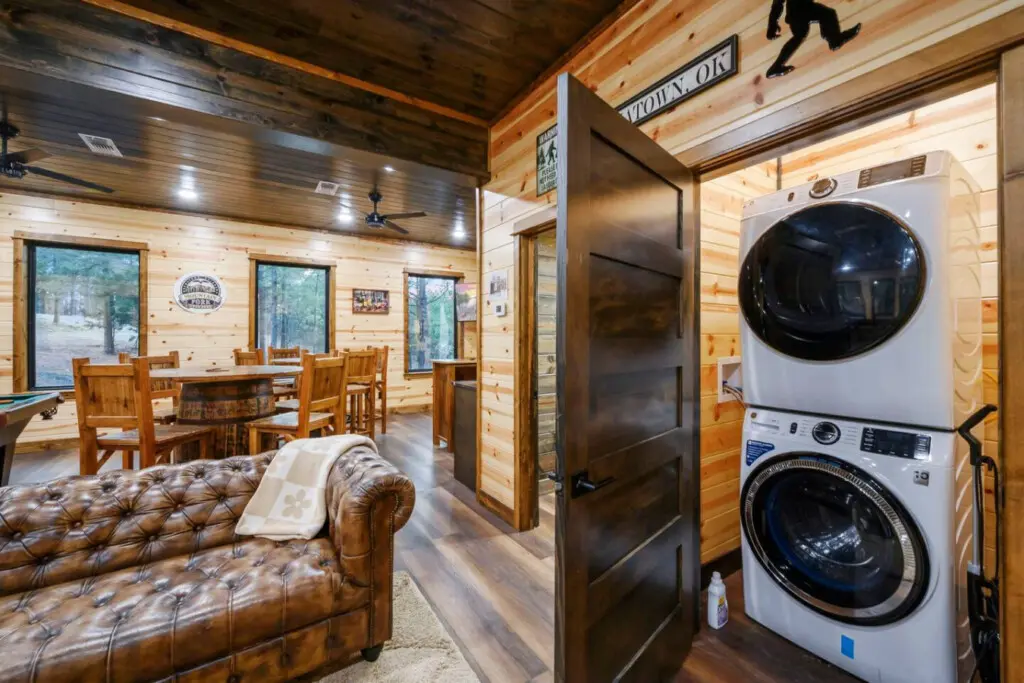 This cozy cabin features rustic wood interiors, a leather sofa, stacked washer/dryer, and beautiful forest views through multiple windows.