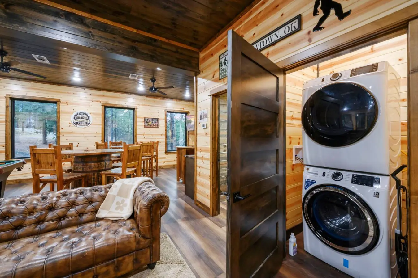 This cozy cabin features rustic wood interiors, a leather sofa, stacked washer/dryer, and beautiful forest views through multiple windows.