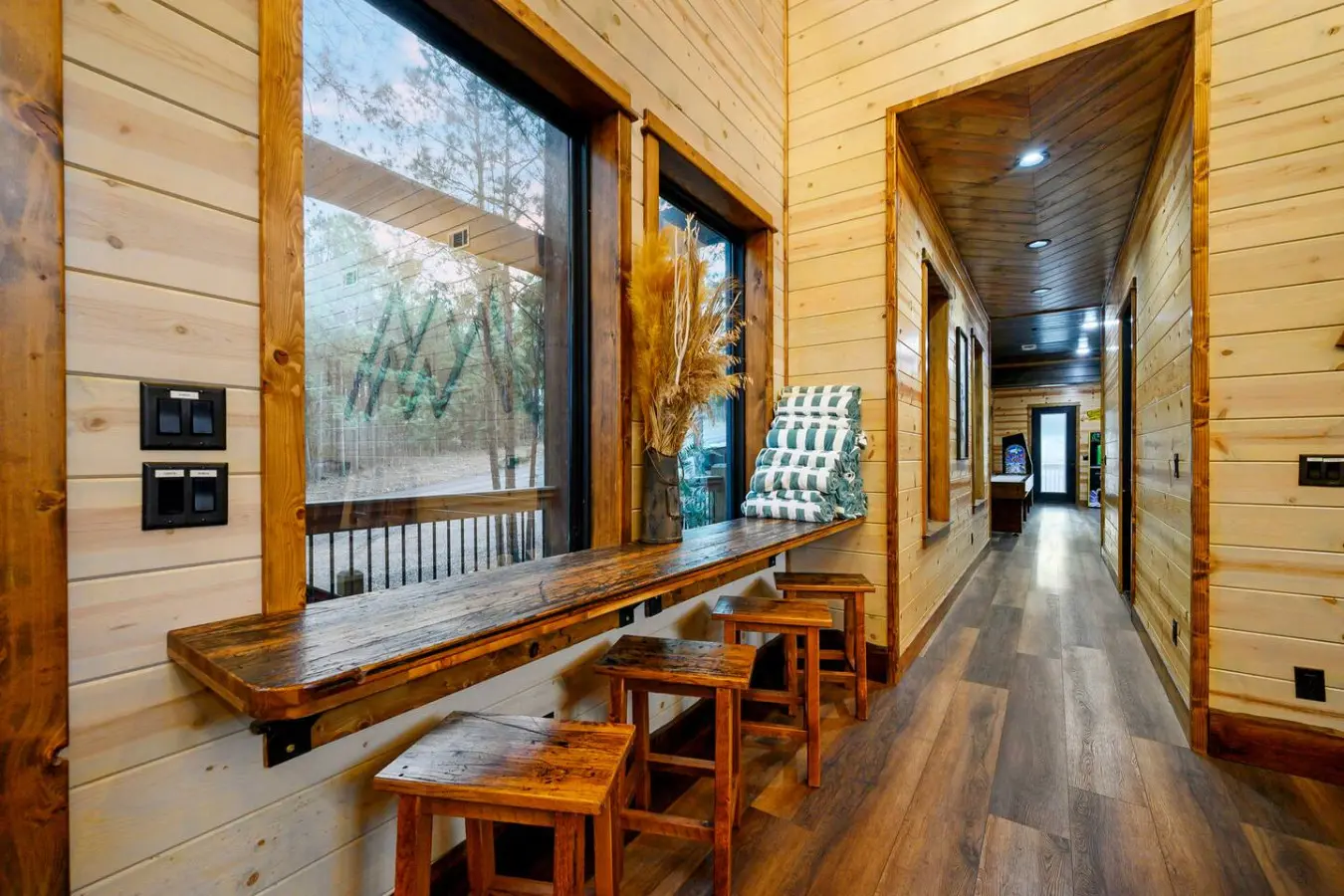This cozy cabin hallway features massive windows overlooking the forest and flows into multiple rooms with beautiful wood finishes throughout.