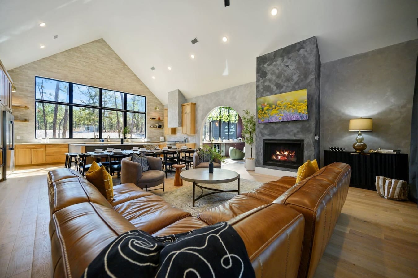 This soaring open-concept living space with its cathedral ceiling, dual leather sofas facing that statement fireplace, and those massive windows framing the forest is basically designed for those moments where you just want to stretch out, get cozy, and l
