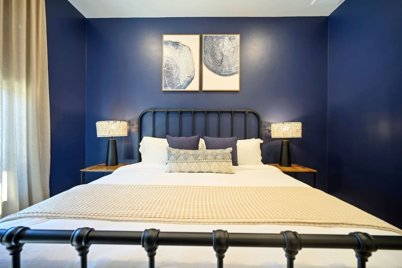 This bedroom is basically the perfect blend of moody sophistication and cozy comfort—deep navy walls, that sleek black metal headboard, warm gold lighting, and those tree-ring art prints overhead make it feel like a stylish forest retreat.