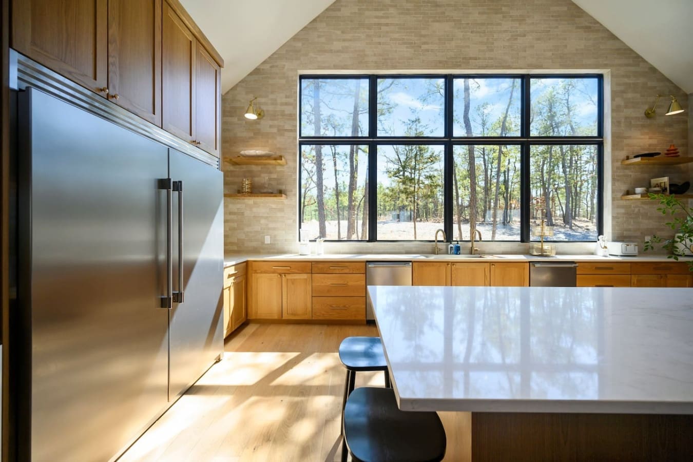 This bright, modern kitchen with its soaring vaulted ceiling and those massive black-framed windows overlooking the forest is basically the ultimate place to cook, hang out, and feel like you're living right in nature without actually roughing it.