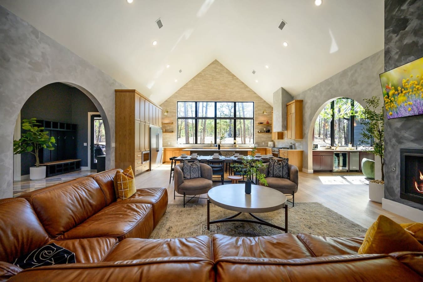 This sprawling great room is basically the ultimate gathering space—soaring ceilings, a stone fireplace, leather sectional, and those massive windows framing the forest that make it feel like you're living right in nature.