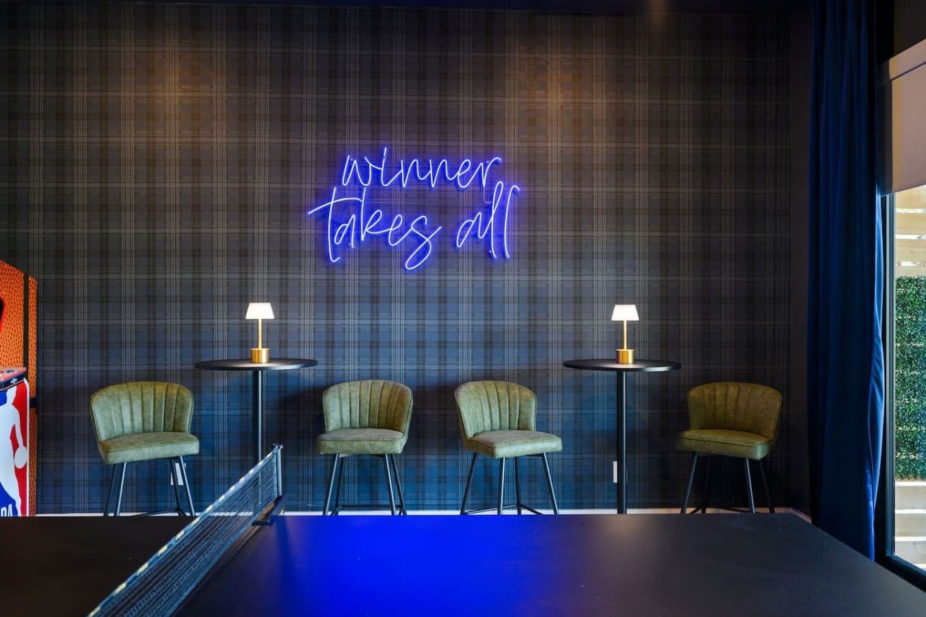 The sleek "winner takes all" neon sign sets a playful vibe above these sage-green velvet chairs—perfect for a little friendly competition or just kicking back in style.