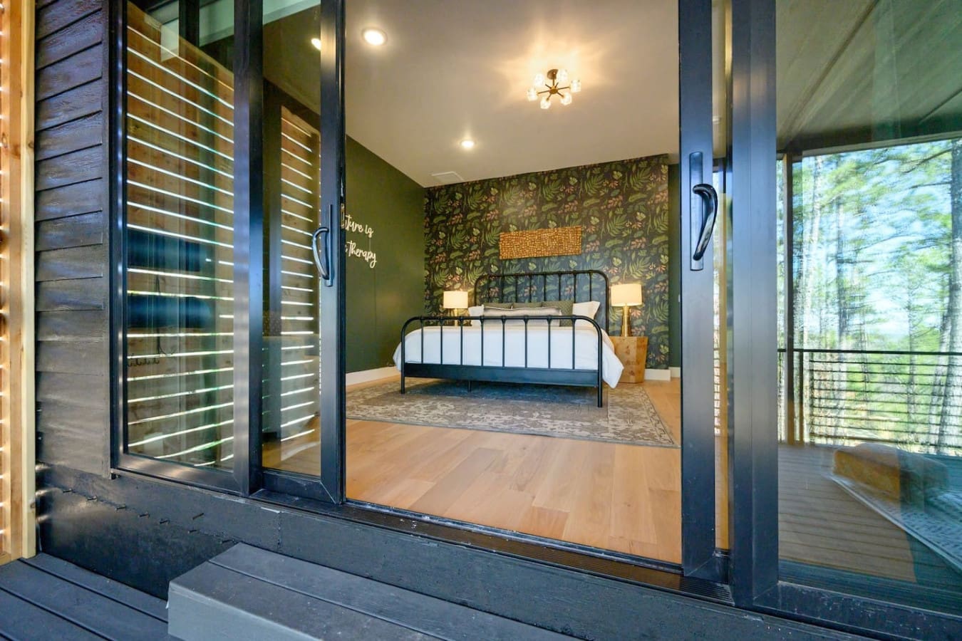 This master bedroom is an absolute dream with those moody patterned walls, a sleek metal bed frame, and those floor-to-ceiling windows that bring the forest right into your space.