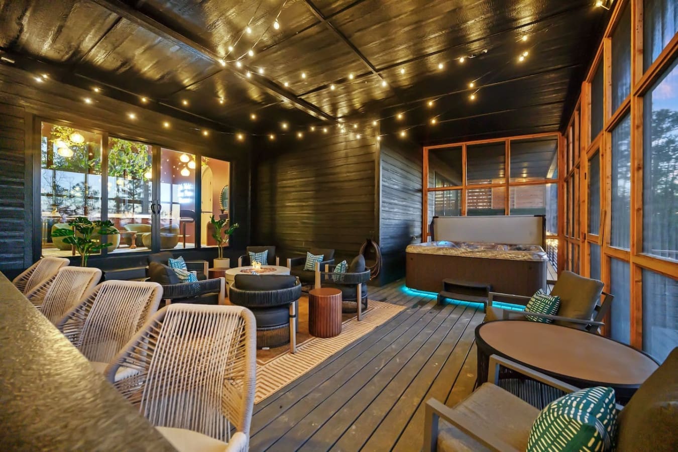 This covered deck is the ultimate hangout spot with those inviting lounge chairs, a cozy fire feature glowing in the corner, string lights twinkling overhead, and those massive windows framing the forest so you've got the best of both worlds—indoor comfor