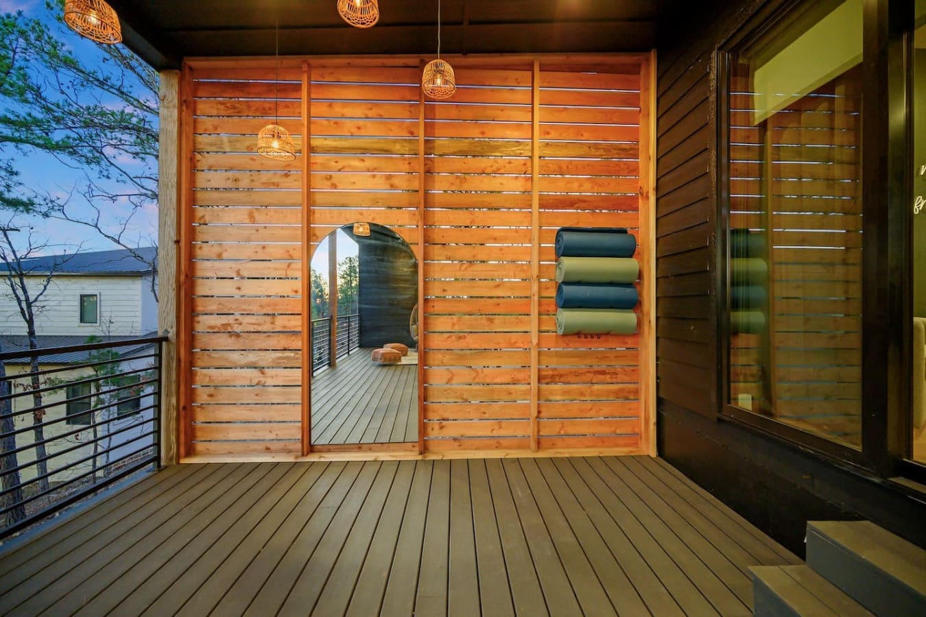 This covered deck with its warm wooden slat walls, hanging pendant lights, and that perfectly framed circular mirror opening onto the forest beyond is basically the ideal spot to step outside without fully stepping away from the cozy vibes.