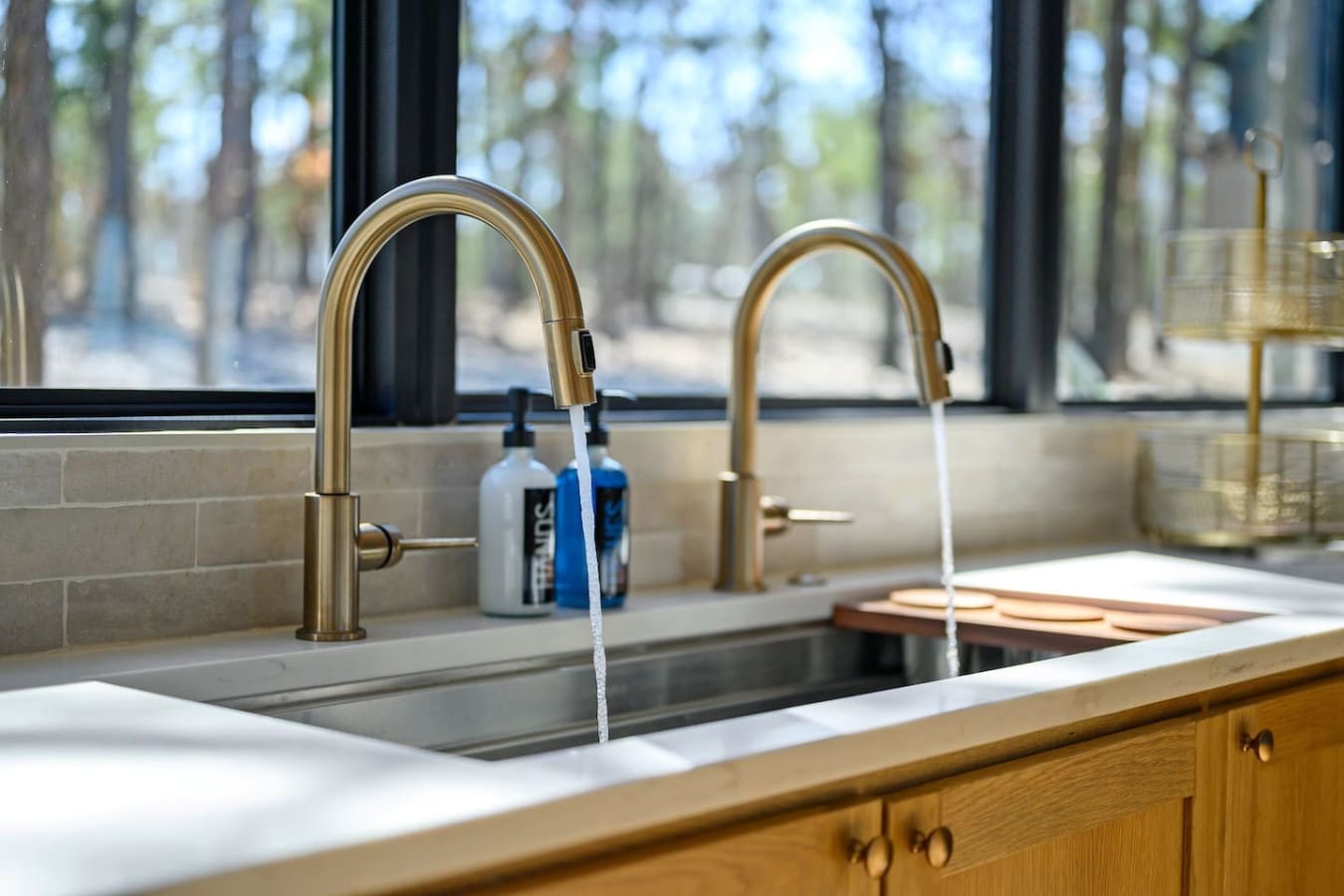The dual bronze faucets with their sleek pulldown sprays and that natural forest view framed perfectly through the window basically make this kitchen sink the kind of spot where you don't mind doing the dishes.