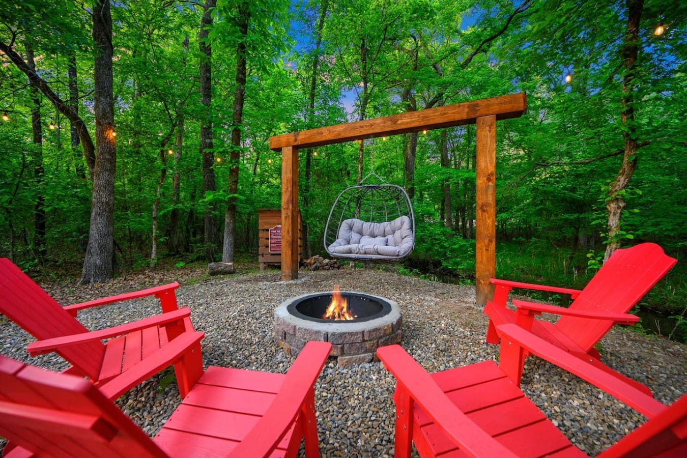 Gather around the fire pit with those iconic red Adirondack chairs, a hanging cocoon swing, and a cozy wooden pergola overhead—basically the ultimate forest hangout spot.