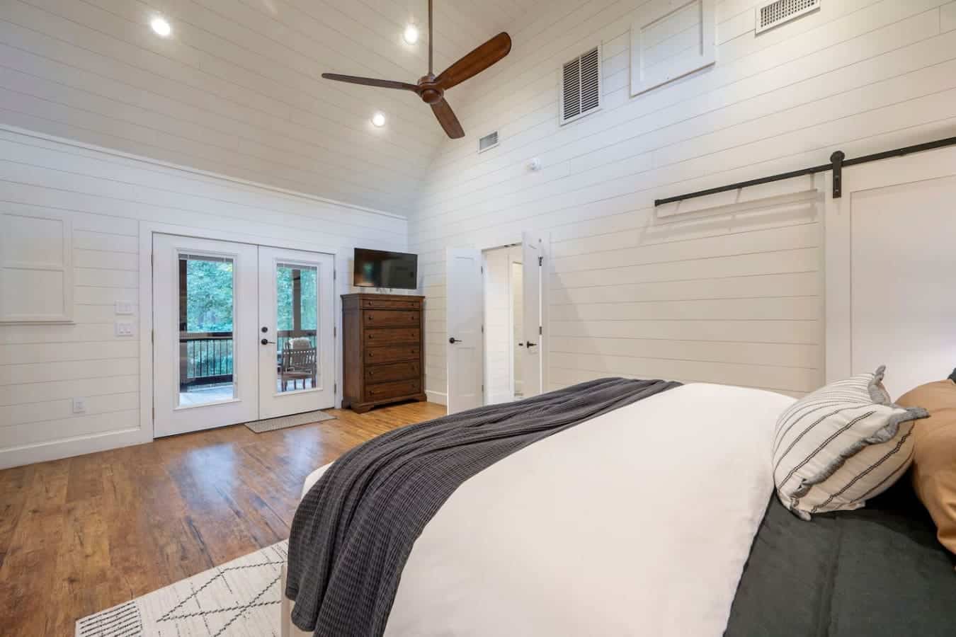This primary bedroom is basically your own private sanctuary with those soaring shiplap walls, a massive bed with room to sprawl, direct deck access, and enough space that you could literally do yoga without stubbing your toe on anything.