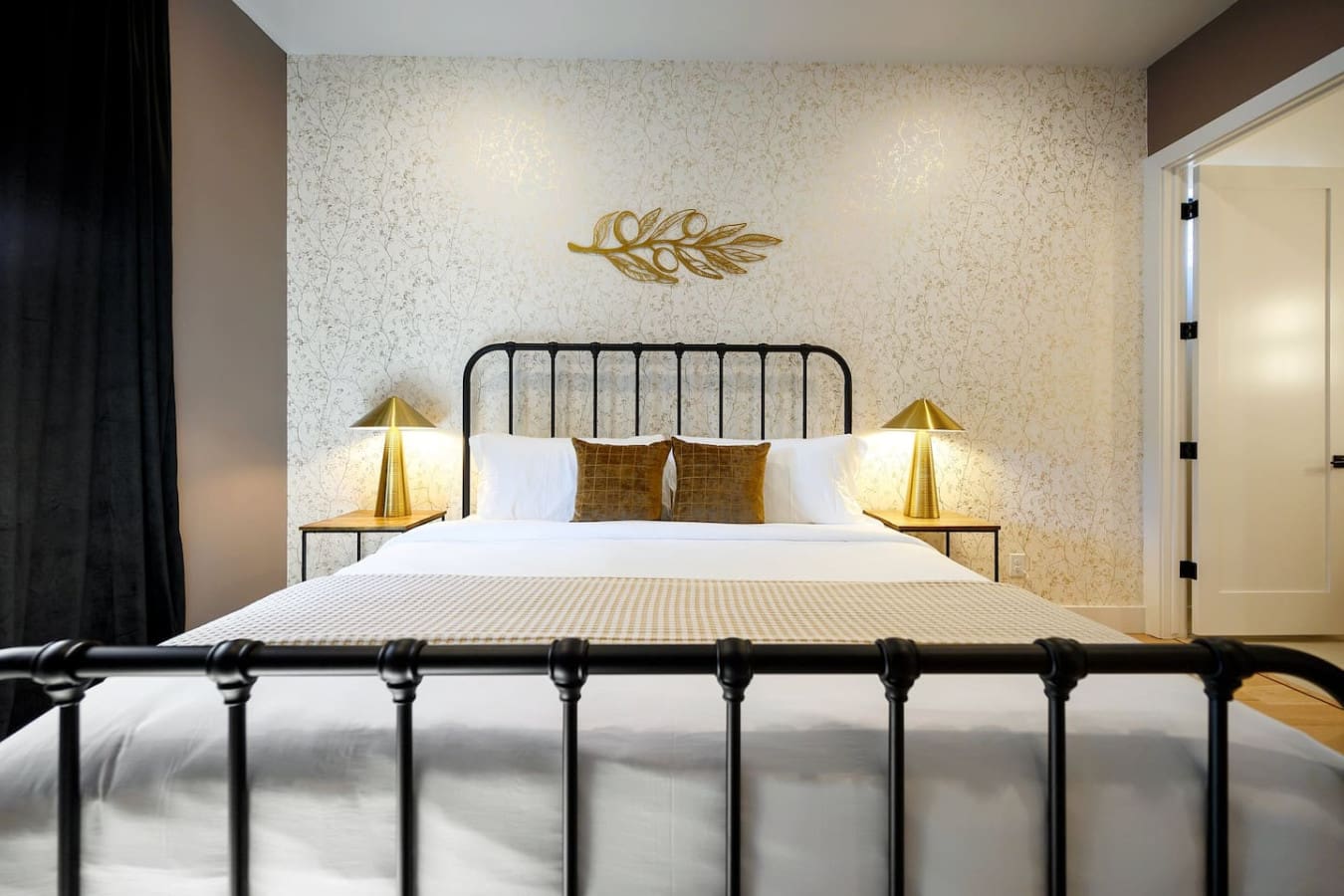 This bedroom is basically the epitome of quiet luxury with that textured cream wallpaper, sleek black metal bed frame, warm brass lamps on either side, and those perfectly placed gold and bronze accent pillows that somehow make the whole space feel both s