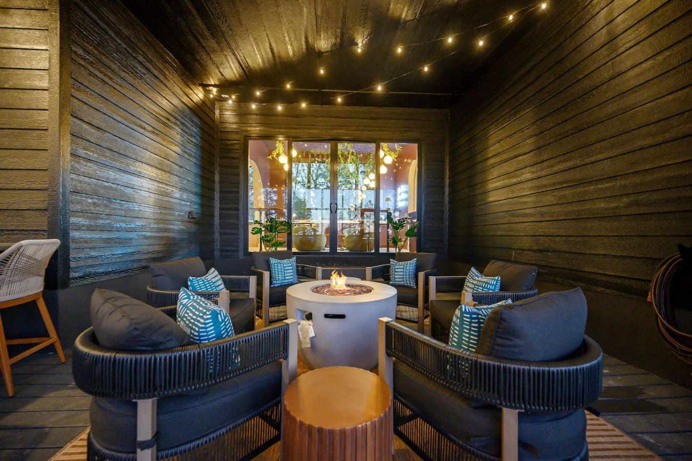 This covered outdoor lounge is basically the perfect spot to curl up by the fire pit with those cozy chairs, string lights twinkling overhead, and those huge windows glowing warm behind you—it's like having your own private treehouse bar without ever leav
