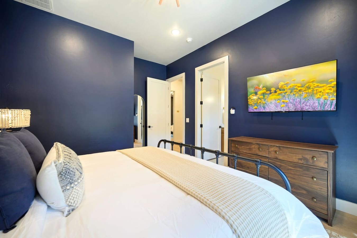 This bedroom totally nails that sophisticated-yet-cozy vibe with the rich navy walls, crisp white bedding, wooden dresser, and that cheerful wildflower landscape art that just makes you want to stay in bed a little longer.