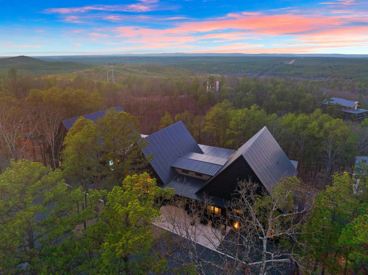 This dramatic modern retreat nestled in the forest with those bold angular roofs and warm glowing windows basically looks like the kind of place where you can watch an epic sunset paint the sky while feeling completely tucked away from the rest of the wor