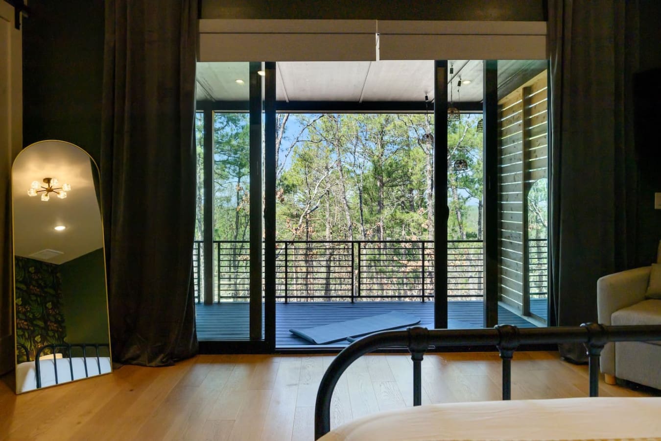 Wake up to floor-to-ceiling forest views from this modern bedroom with its sleek black bed frame, warm wood floors, and that stunning blue deck stretching out like your own private treehouse escape.