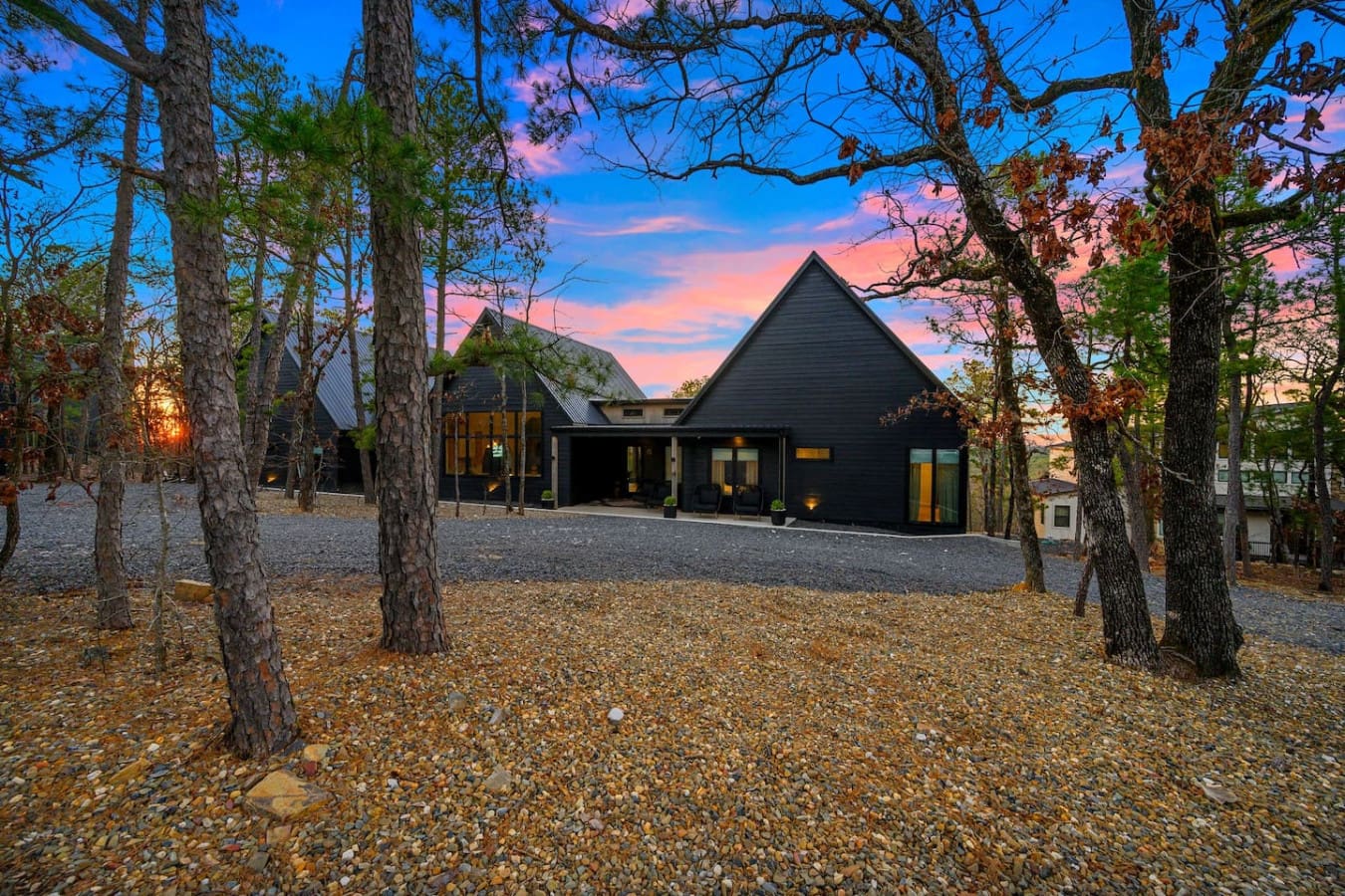 Picture this: a stunning modern retreat nestled among towering pines with those dramatic peaked roofs and warm glowing windows, basically looking like the kind of architectural escape where you can watch that epic sunset paint the sky while feeling comple