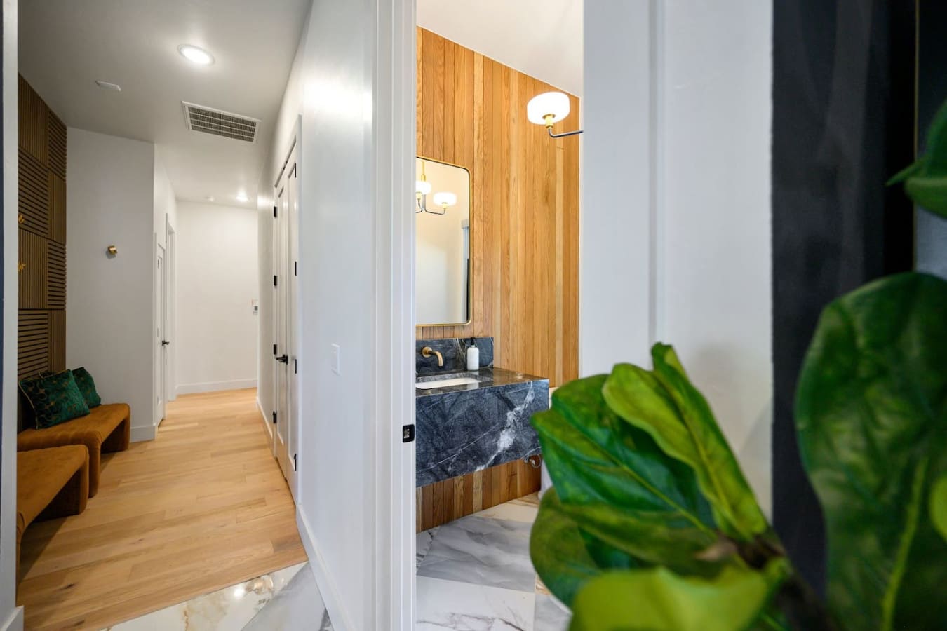 The hallway and bathroom space comes together with that warm wood accent wall, sleek dark marble countertop, and bright modern lighting that somehow manages to feel both spa-like and super welcoming.