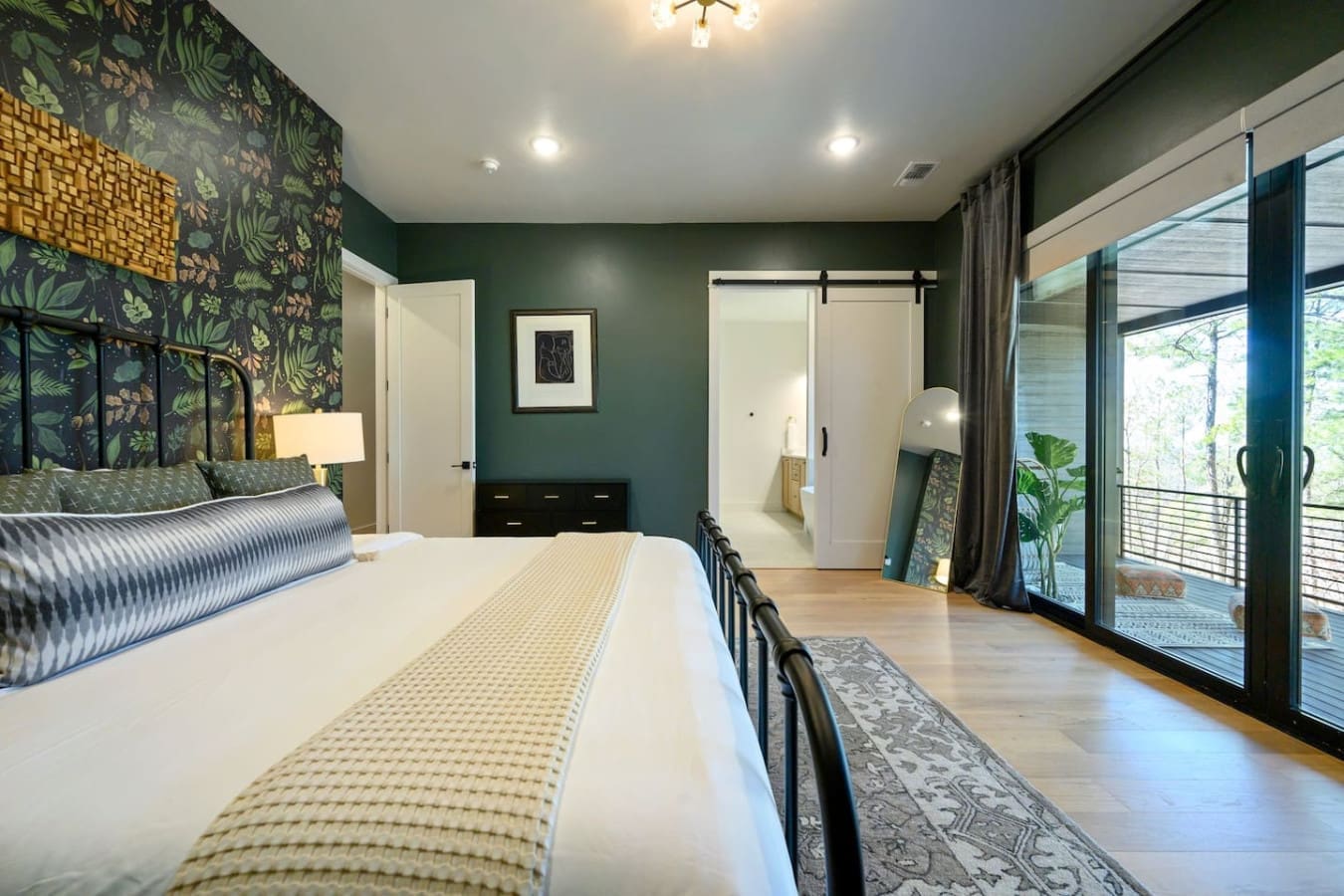 This master bedroom is basically giving you all the luxury forest-retreat vibes with that rich green accent wall, statement botanical wallpaper, sleek black metal bed, and those massive sliding glass doors that open right onto the deck so you can wake up