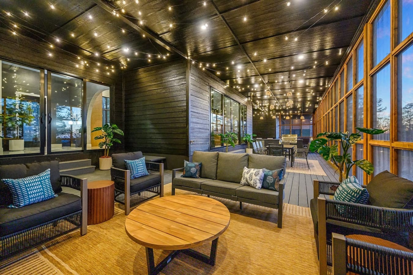 This covered deck with those glowing string lights, cozy seating nooks, and floor-to-ceiling windows basically feels like your own private treehouse lounge where you can curl up and watch the forest come alive at dusk.
